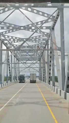 The video shows a view from the driver's seat of a vehicle moving across a bridge. The bridge has a large, gray metal truss structure overhead with circular cutouts. The road has two lanes, marked by a solid white line and a yellow line. There are black and white striped barriers on the side of the road. Several trucks are visible on the bridge, with one in the foreground on the left and another further ahead in the right lane. The sky is overcast.