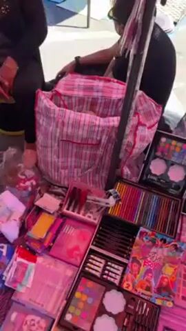 The video shows people shopping at an outdoor flea market on a sunny day. Some tables are visible with various products on display. One table is covered with a pink tablecloth and filled with toys and accessories, including lip balms, figurines, dolls, and makeup sets. Another table is covered with a white cloth and displays miniature Disney characters. There are people standing behind the tables, presumably vendors, and customers examining the goods. There are cardboard boxes behind the tables and people walking in the background. The atmosphere is bustling.