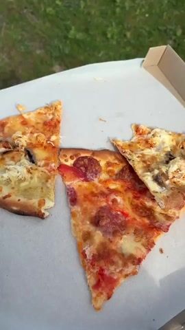 The video starts with a close up of a pizza box with three slices of pizza inside. One slice is pepperoni, and the other two appear to have cheese and mushrooms. The camera then pans up to reveal a view of a green field with a road and power lines in the distance. The sky is blue with white clouds. The camera then pans back down to the pizza box, which is now in the foreground. The view of the mountains is visible in the background.