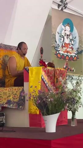 A man in a yellow robe sits cross-legged on a stage and reads from a paper. He has a microphone headset on. He is behind a table draped with yellow and red cloth and decorated with two vases of flowers. Behind him is a tapestry depicting a white, multi-armed figure seated on a lotus. The stage is red. The room is white.