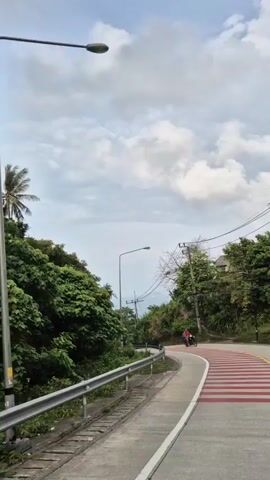 The video shows a winding road with red and white striped pedestrian crossing. A person on a scooter drives on the road, past the camera. Green foliage flanks either side of the road. The sky is overcast with fluffy clouds. In the distance, glimpses of the ocean are visible through gaps in the trees. Utility poles and streetlights line the road. The video is shot from a stationary position.