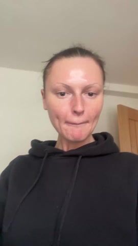 The video shows a woman with her hair pulled back, wearing a black hoodie. She smiles slightly, then raises her hand to her head. She then looks up and speaks. The background is a plain, light-colored wall with a door frame visible on the right.