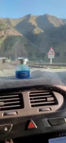 The video shows a view from inside a car, looking forward. The dashboard is visible in the foreground with air vents and a fuzzy grey mat covering part of it. On the mat, a small, blue bottle of car air freshener with a silver cap is prominently placed. The car is on a highway, with other vehicles, including trucks and cars, visible in the distance. The road curves slightly to the left. On either side of the highway are steep, terraced hillsides covered in dry vegetation and sparse trees. The sky is clear and blue, and the sun appears to be low in the sky, casting long shadows. The overall impression is that the car is driving through a mountainous, arid landscape.