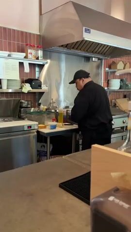 Here is a detailed description of the video:

The video shows the inside of a kitchen, likely in a restaurant. A person wearing a black uniform and a baseball cap is cooking on a flat top grill. The person moves between a small work station and the grill. On the grill, there is bacon, potatoes, and a sausage or hotdog. The work station has multiple bottles and containers on it, including condiments and cooking ingredients. Behind the grill is a stainless steel hood and a red tiled backsplash. There are shelves above the work station with kitchen supplies and plates. In the foreground is a counter.
