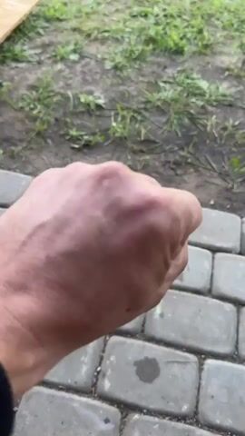 A close-up shot shows a Caucasian male's right hand. The hand is clenched into a fist, then slowly opens to reveal a middle finger extended. The hand is dirty and appears weathered. The background is blurred, showing a patch of grass and a brick walkway. The lighting suggests it is daytime.