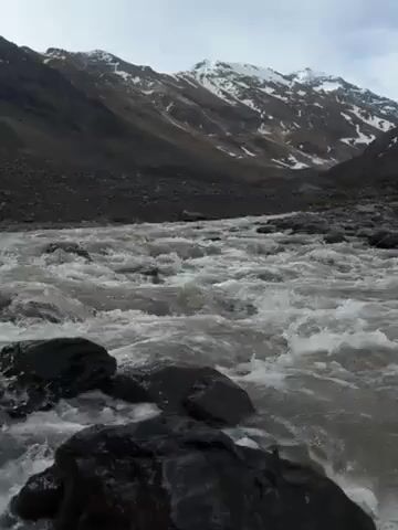 The video shows a fast-flowing river surrounded by mountains. The river water is turbulent and mostly white, flowing over dark rocks. The mountains in the background are rugged, with patches of snow on their peaks and dark grey or brown slopes. The sky is overcast.