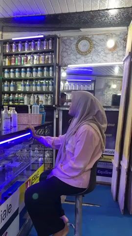 The media file captures an interior scene, likely a shop, with a focus on shelves filled with numerous bottles. A man, wearing a black t-shirt, stands behind a counter, appearing to be attending to the bottles. He is positioned to the left of the frame. To the right, a woman wearing a light pink top and a light purple hijab is seated on a stool, facing the counter. She is looking towards the man and the counter. The counter itself is made of glass and has a blue light strip running along its edge. Behind the counter, more bottles are visible, as well as a patterned wall. Above the shelves of bottles, a white ceiling with lighting fixtures is visible, and a blue light also illuminates the top of the shelves. A circular decorative element with Arabic script is mounted on the wall. A "STOP!" sign with a prohibition symbol is prominently displayed on the front of the counter. The overall impression is that of a retail establishment selling various liquid products, possibly perfumes or essential oils, given the large number of similarly shaped bottles and the "essenti" text visible on a label. The lighting suggests it might be evening or an indoor setting where artificial lights are in use. The presence of a customer and a shopkeeper indicates a commercial transaction or interaction is occurring.