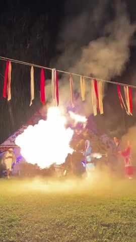 This video shows a nighttime outdoor performance. In the foreground, a person in a blue costume is holding a long pole that is emitting a large burst of fire and smoke. To the right of this person, two other individuals in traditional attire are holding a similar pole, possibly assisting with the fire effect. In the background, there appears to be a tent-like structure and a large, decorated object, possibly a dragon effigy, also illuminated by the fire. Red and white streamers hang from a wire overhead, fluttering in the heat and smoke. The ground is covered in grass, and the overall atmosphere is dramatic and festive.