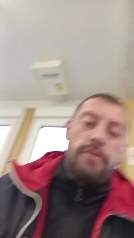 The video shows a man holding up a gray, metallic ring towards the camera. He is wearing a black jacket with a red hood. The ring is initially oriented vertically then the man rotates it, showing the circular opening within the ring. A white ceiling with a light fixture and electrical panel is visible in the background. The man looks at the camera throughout.