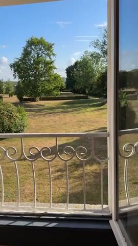 The video opens with a view through an open door or window to a sunny landscape with trees and a manicured lawn. A white decorative metal railing is visible in the foreground. The camera then pans to the right, revealing an interior space. There is a wooden dresser with a glass top and a decorative lamp on it. To the left of the dresser is a vintage armchair upholstered in a plaid fabric. A decorative mirror hangs on the wall above the dresser. The camera continues to pan, showing more of the interior and the view out the window. The window appears to be a French door leading to a balcony. The interior walls are painted a light color with a dark-colored wainscoting. The floor is made of wood planks. The video ends with a closer view of the window and balcony.