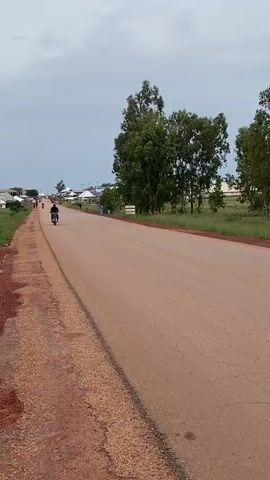 A person rides a motorcycle down a dirt road. A dark-colored sedan passes the motorcycle on the road. The road is lined with green grass and trees. There are buildings in the distance. The sky is overcast.