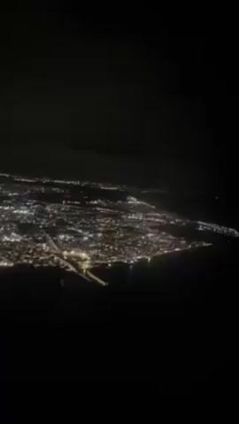 The video depicts a night aerial view of a city with a network of lights illuminating the landscape below a dark sky. Some clouds are visible above the urban area. In the foreground, the distinct pattern of airport runway lights is evident.
