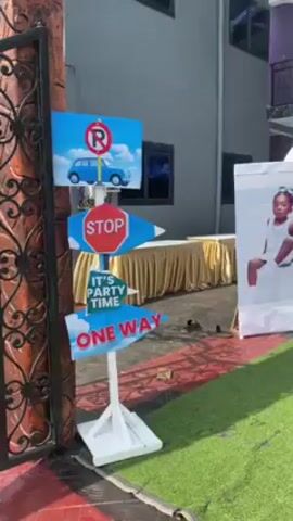 The video depicts a decorated outdoor setting for a birthday party, likely a child's. There is a signpost featuring road signs and the text "It's Party Time," as well as large freestanding posters of a young child, presumably the birthday celebrant. Tables with white linens and place settings are arranged under an arch decorated with blue and white balloons. A banner reads "Welcome to Joel-Ethan Birthday Party." The setting is partially covered with artificial turf and is surrounded by a building. People are seen moving in the background.