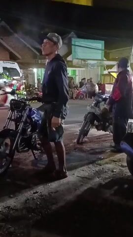 A man wearing a cap, black hoodie, and shorts stands next to a blue motorcycle at night. He appears to be holding a cigarette in his mouth and looking upwards. Another person in a dark jacket with a design on the back stands near a grey motorcycle to the right. Several other people are visible in the background, some near motorcycles, in what appears to be a street or open area with buildings and lights.