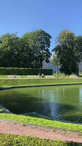 The video shows a bright, sunny day with a clear blue sky. In the foreground, there is a vibrant green pond or lake with ripples on its surface. A dirt path with scattered gravel runs along the edge of the pond, bordered by green grass and low hedges. Beyond the pond, a lush green lawn stretches towards a white picket fence that delineates a walkway. Several people are walking along this walkway, some appearing to be individuals and others in small groups. Behind the walkway and fence, tall, mature trees with dense green foliage create a shaded backdrop. In the distance, a large, ornate blue and white building, possibly a palace or grand estate, is visible through the trees. The architecture of the building suggests a European style with multiple windows and a structured facade. The overall scene depicts a peaceful and scenic park or garden setting.