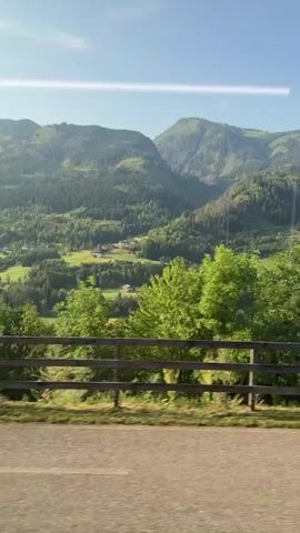 The video shows a landscape view from a moving vehicle, likely a car or train. The foreground features a paved road with yellow lines, suggesting a highway or scenic route. A wooden fence runs along the side of the road, separating it from a grassy area and trees. In the middle ground, rolling green hills dotted with trees and some scattered houses are visible. The background is dominated by majestic, lush green mountains under a clear blue sky. The sun is shining, casting a bright light and creating a lens flare effect in the upper left corner of the frame. The overall impression is one of a picturesque rural or mountainous region during daytime.