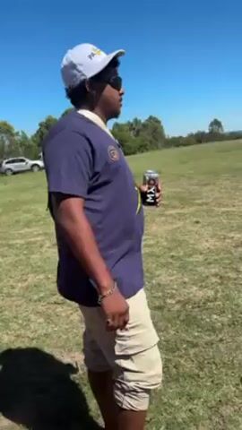 A medium shot shows a man standing outside on a sunny day, drinking from a black and white can. The man has dark curly hair and is wearing sunglasses, a white baseball cap with text, a dark blue short-sleeved shirt, and tan cargo shorts. He also has a chain bracelet on his left wrist.  He drinks from the can and then lowers it, looking down at the can. The ground is covered in dry grass, and in the distance are trees and a parked car. Loud music is playing.