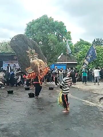 The video shows a street performance in a rural setting. In the center of the frame, a performer wearing a large, elaborate mask with a lion-like face and a tall, dark headdress is the focal point. The performer is dressed in traditional attire, and their movements suggest a dance or ritual. To the right of the mask performer, another person in a striped shirt and pants holds a musical instrument, possibly a drum or tambourine. Several other people in similar striped outfits are gathered around, some appearing to dance or move in sync with the performance. A crowd of onlookers is visible in the background, watching the event. In the background there is a building, trees, and banners. The ground is asphalt, and the sky is overcast, suggesting it might be a cloudy day.