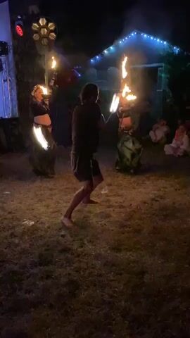 This is a nighttime video of an outdoor performance involving fire. In the foreground, a barefoot individual wearing a dark t-shirt and shorts is actively spinning fiery objects, likely poi, which are creating bright streaks of flame. To the left and right in the midground, two individuals, appearing to be women, are also holding and manipulating flaming props. The person on the left is wearing a dark top and a dark, flowy skirt. The person on the right is wearing a dark top and a green, flowy skirt. Both are adorned with what appear to be headdresses. In the background, there is a structure decorated with string lights and a stage-like area with lighting equipment, including a large, circular light fixture. Several spectators are seated or standing in the background on the right side of the frame, partially illuminated by the performance. The ground appears to be grass or dirt, with scattered debris. The overall atmosphere suggests a performance or festival event.