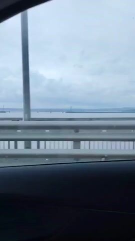 The video was filmed from inside a moving vehicle traveling across a bridge. Through the passenger side window, a grey metal guardrail is visible. The vehicle is moving fast enough that the vertical posts along the rail blur as they pass by. Beyond the rail is a large body of water, with a second bridge spanning it in the distance. This second bridge has towers and appears to be a railroad bridge.  The sky is overcast, with dense clouds. The vehicle's interior is dark, only the dashboard and window trim can be seen. The sound of music can be heard in the background.