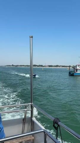 The video captures a boat trip on a sunny day. The camera is positioned on a boat, looking out over the water. In the foreground, the stainless steel railings of the boat's deck are visible. 

In the midground, another, smaller, blue boat speeds across the water, leaving a white wake behind it. There appear to be three people aboard. To the right of the small boat, a larger, blue and white boat is docked or anchored. In the distance, a coastline is visible under a clear blue sky. The water is a turquoise color, and there are some small waves creating a ripple effect. The overall scene depicts a typical day on the water with various types of boats in use.