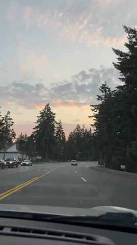 The video is captured from inside a vehicle driving on a road bordered by tall evergreen trees. The sky displays a soft sunset with clouds tinged with pink and orange hues. Several cars are visible on the road, moving in the same direction as the camera. A building with a white exterior and dark roof is situated on the left side of the road, and a traffic light is visible ahead. The overall scene depicts a tranquil drive during sunset.