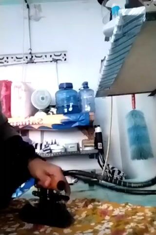 A person in a dark headscarf and clothing is ironing clothes on a patterned ironing board. A blue water cooler jug and a smaller plastic bottle of water are visible on a shelf behind the person. A white oscillating fan is also on the shelf. A light blue duster hangs from the ceiling on the right side of the frame. The room appears to be a domestic space with white walls and simple shelving. The person is holding the iron with their right hand and appears to be moving it across the fabric. The timestamp indicates the video is a short clip of this activity.