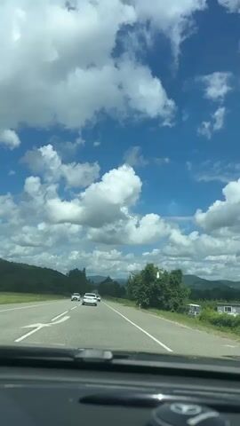 This is a first-person view from inside a vehicle traveling on a paved road under a blue sky with scattered white clouds. The road has multiple lanes and white markings. Several vehicles are visible on the road ahead, including at least three cars and one SUV with a roof rack. The landscape on either side of the road consists of green trees and hills, with some hints of distant mountains. The sun appears to be shining, casting light on the scene. The interior of the vehicle is partially visible in the foreground, including the dashboard and a circular object with a logo. The video captures a sense of motion as the vehicle travels along the highway.