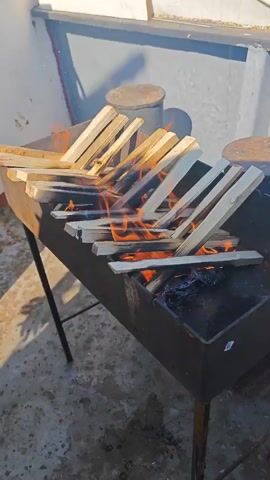 A charcoal grill is set up on a surface that appears to be a deck. Pieces of wood are arranged in a V-shape over the grill, with flames visible underneath. The wood is beginning to char and smoke. In the background, there are cylindrical objects that could be bollards, and a body of water is visible, suggesting the location is near or on the water. The lighting suggests it is daytime.