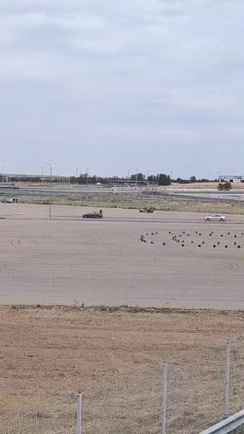A wide shot of a large, flat, paved area with some sparse vegetation in the foreground and a fence. Several vehicles are visible on the paved area, along with what appear to be traffic cones forming a course. In the middle ground, a dark-colored sedan is driving and appears to be drifting or spinning. A yellow vehicle is also visible further back, and two people are standing near it. In the background, there are more paved areas, some structures, and a cloudy sky.