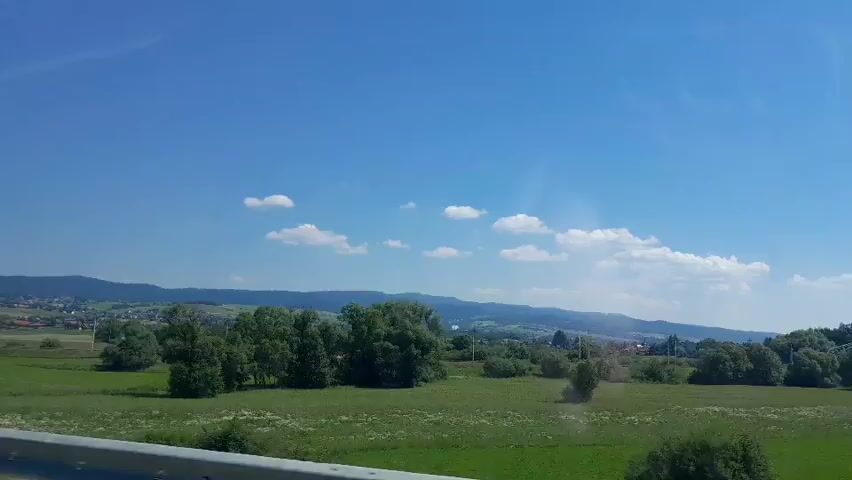 The video shows a scenic view of a rural landscape on a clear day. The sky is a vibrant blue with scattered white clouds. In the foreground, a guardrail runs horizontally across the bottom of the frame. Beyond the guardrail, a lush green field stretches out, dotted with several trees and bushes. In the distance, rolling hills and mountains form the horizon, with a small village or town visible nestled among them. The overall impression is one of peacefulness and natural beauty. The camera appears to be in motion, suggesting that the viewer is traveling through the landscape, possibly by car or train.
