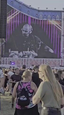 A large screen displays a bald DJ wearing headphones and a dark shirt, with purple lights in the background. Below the screen, a crowd of people stands in a grassy outdoor area, some dancing. The overall environment appears to be a music festival or concert.