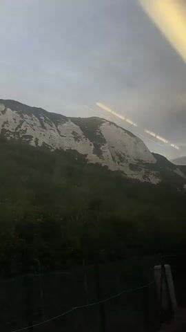 The video shows a view from a moving vehicle, likely a train, looking out a window at a large, white-streaked cliff face covered in green vegetation. The sky is overcast with a greyish hue. The cliff appears to be made of chalk or a similar material, with patches of white exposed rock. Trees and bushes cover the lower slopes of the cliff. The vehicle's interior lights are visible as streaks of yellow and white across the top of the frame, indicating motion blur. A dark fence is visible in the foreground, close to the window. The overall impression is a scenic but somewhat gloomy landscape.