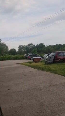 The video presents a panning view of an outdoor, semi-rural area, likely a private yard or informal parking space, with several cars parked on grass next to a concrete strip. The setting includes a backdrop of green, tree-covered hills under an overcast sky, suggesting a cloudy day.

The camera first shows a red minivan and a dark car, with a wooden box (possibly a beehive) in the foreground. It then pans to a blue Volvo XC60 SUV (license plate "CT 8586 TH"). Following this, a silver BMW (license plate "CT 8034 PX") and a dark Peugeot sedan (license plate "C 6440 HC") are visible, with a utility pole in the background. The pan continues to a green Fiat Panda hatchback (license plate "CT 5840 AP") parked with its rear facing the camera, near a blue tarp. The final shot reveals a pile of wooden debris and general clutter on the grassy ground, extending into the distant green landscape.

No people are visible in the video. The visible license plates are Bulgarian, consistent with the location context of Buzovgrad, Bulgaria.