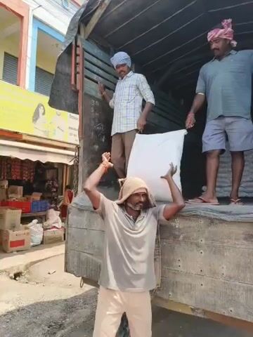 The video shows three men unloading sacks from the back of a truck. One man is inside the truck, assisting with the unloading process. Another man is standing on the edge of the truck bed, holding a sack that he is passing to a third man who is carrying it on his head and shoulders. The third man is walking away from the truck, presumably to unload the sack. The scene appears to be outdoors, with a building in the background that has a sign in a foreign language. There are stacks of sacks visible on the ground next to the truck, suggesting that this is a delivery or distribution point. The men are dressed in casual clothing, and the overall atmosphere suggests manual labor.