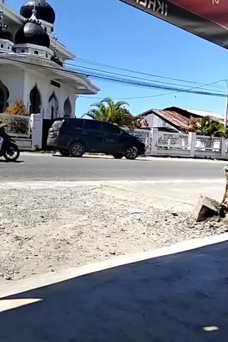 The video shows a view of a street with a black car and a motorcycle driving by. The motorcycle has two people on it, with the passenger wearing a white headscarf and a red shirt. A mosque with multiple black domes is visible in the background to the left of the car. Other buildings and foliage are across the street as well, behind a fence. The sky is clear and blue. The camera appears to be stationary, shooting from under a red sign.