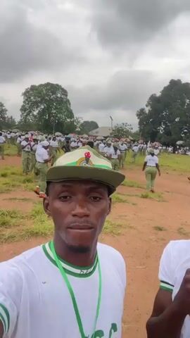 The video begins with a close-up selfie of a young Black man wearing a white t-shirt with green trim and a matching green and white cap. He has a serious expression, and the background shows a blurred outdoor scene. As the video progresses, the camera pans slightly, revealing another young Black man to his right, also wearing a similar uniform and holding his hand to his mouth. The background becomes clearer, showing a large group of people, mostly young Black individuals dressed in white t-shirts and green pants, wearing similar green and white caps. They are gathered in an open, grassy field with some trees and tents visible in the distance. Some people are holding umbrellas. The two men in the foreground continue to pose for the camera, with the man on the left making a kissing gesture and sticking out his tongue, and the man on the right flashing a peace sign. The camera then zooms out slightly, showing more of the crowd in the background, some of whom appear to be walking or standing in organized groups. The overall impression is of a large gathering or event, possibly a ceremony or training camp, where participants are dressed in a uniform. The lighting suggests an overcast day.