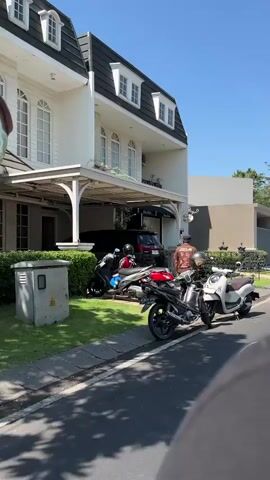 A man wearing a brown t-shirt and black shorts stands next to several parked motorcycles and scooters on a paved street in front of a large, white house with black trim and dormer windows. The house has a covered carport with a dark-colored car parked underneath. The motorcycles and scooters are lined up along the curb. The man appears to be looking at the vehicles. The sky is clear and blue. Sunlight casts shadows on the street and the house.