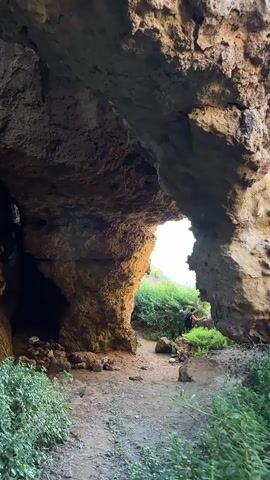 The video displays an interior view of a natural rock cave, looking outward through a large, irregular opening. A dirt path extends from the foreground towards the exit, flanked by scattered rocks and small green plants. Outside the cave, green bushes are prominent, and a person wearing dark clothing stands near a large rock on the left side of the cave's opening, facing towards the sea. As the camera moves forward, the view through the opening becomes clearer, revealing a calm body of water, likely the sea, extending to the horizon. In the distance, a distinct, pointed landmass is visible over the water, framed by the cave's rocky outline.