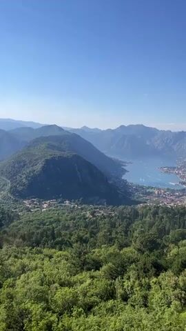 The video shows a panoramic view of a bay surrounded by mountains. The bay is calm and blue, with a town visible on its shores. The mountains are covered in lush green vegetation, and the sky is clear and blue. The camera appears to be moving slightly, panning across the landscape. The overall impression is one of natural beauty and tranquility.