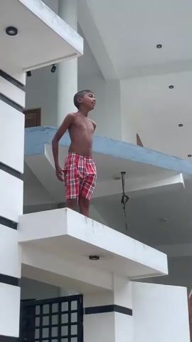 A young boy, shirtless and wearing red and white checkered shorts, stands on a white balcony. He is looking to his right and smiling. Behind him, the sky is overcast, suggesting it could be daytime or evening. The boy appears to be in a residential area, with parts of a house and a decorative balcony visible. The architecture suggests a tropical location, possibly in Sri Lanka, given the contextual information. The boy's posture and expression do not indicate any distress or particular urgency. He is the sole subject in the frame.