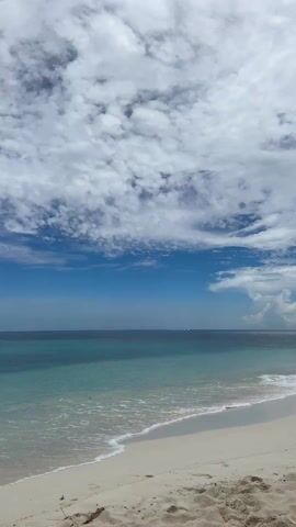 The video shows a white sand beach on a sunny day with a partly cloudy sky. The water is turquoise near the shore, transitioning to a darker blue further out. Small waves gently lap the beach. Two small boats are visible in the distance on the horizon. The camera tilts upward towards the sky.