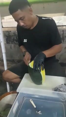 A man wearing a black t-shirt and black gloves is sitting and cutting a yellow watermelon with a knife. He is using a white cutting board. There is a clear plastic container in front of him with a spoon inside. In the background, there are white boxes and a white pole. The man is focused on cutting the watermelon.