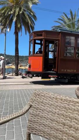 A tram passes to the left of the camera's viewpoint. The tram is brown with an orange section at both ends and the words "FC. DE SOLLER S.A." on the side. Passengers sit in the open section of the tram, waving to the camera. The tram moves on cobblestone tracks. The background features palm trees, a clear blue sky, and a glimpse of a harbor. A wicker chair is prominently in the foreground, adding to the scene's ambiance.