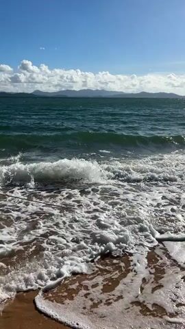 The video captures a dynamic view of the ocean with waves crashing on a sandy shore. The water is a deep teal color, and white foam from the waves covers the foreground. In the distance, a hazy mountain range is visible under a bright blue sky dotted with fluffy white clouds. The sun casts a warm light, creating a sense of a pleasant day. The camera appears to be positioned low, near the water's edge, giving the viewer a sense of being present at the scene. There is a thin dark rope or line running horizontally across the lower left portion of the frame. In the upper right quadrant of the video, a distant figure on a board, possibly windsurfing or kiteboarding, can be seen against the horizon.