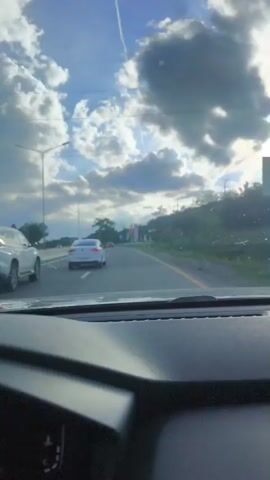 The video shows a first-person perspective from inside a moving car, driving on a multi-lane highway under a partly cloudy sky. The car is on the right side of the road, and other vehicles, including cars and a motorcycle, are visible in front and to the left. The camera pans slightly to the right, revealing a large monument or sign on the roadside that reads "KION VOYAGE." The surrounding environment includes trees, streetlights, and concrete barriers.
