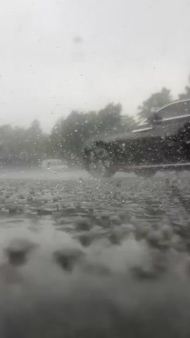 A car drives through a downpour on a road in a misty, outdoor setting. The ground is wet and reflects the surroundings. The sky is overcast, and trees are visible in the background, partially obscured by the rain and mist. The overall atmosphere is gloomy and wet, suggesting a heavy rain shower. The car's tires are kicking up water as it moves. No people are visible in the frame, only the car and the natural elements of a rainy day. The image's low angle gives a sense of being close to the ground, emphasizing the intensity of the rain. The time of day is ambiguous due to the overcast conditions, but it appears to be daytime. There is no visible text in the media file.