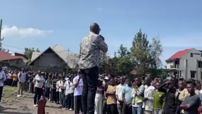 Here is a description of the video:

A man stands on a low platform, addressing a large crowd of people. He is wearing a patterned shirt, dark pants, and sunglasses. He holds a microphone in his right hand and gestures with his left. The crowd is predominantly male, and they are listening attentively to the speaker. The background includes buildings with varying architectural styles, trees, and a blue sky with some clouds. The setting appears to be an outdoor gathering, possibly a rally or a public address in a populated area.