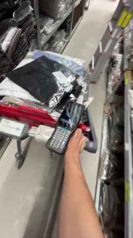 The video shows a first-person perspective of someone pushing a shopping cart filled with clothing items through a warehouse or stockroom. The shelves are packed with more merchandise, mostly apparel in clear plastic packaging, stacked high. A silver ladder is visible on the right side of the aisle, suggesting items may be stored at a considerable height.

The person is holding a handheld scanner device, which appears to be a barcode scanner, and the screen is displaying some information. The scanner is black with a keypad and a screen that shows a grid-like interface with some text and graphics. The cart itself is red and grey and has a "Carrefour" logo visible on the front, indicating it's likely a shopping cart from the Carrefour supermarket chain, which operates in Israel.

The clothing items on the cart include at least one black garment in a plastic bag, and several other items of clothing in various neutral colors, also packaged. Some of the packaging has tags attached.

The overall scene is utilitarian and suggests an inventory or stocking activity. The lighting appears to be artificial, consistent with an indoor warehouse environment. There are no clear indicators of the time of day or weather conditions. The focus is on the task at hand, with no discernible emotions expressed. The text "Carrefour" is clearly visible on the cart. Other text on packaging is too small or blurry to be read. The location is identified as Holon, Israel.