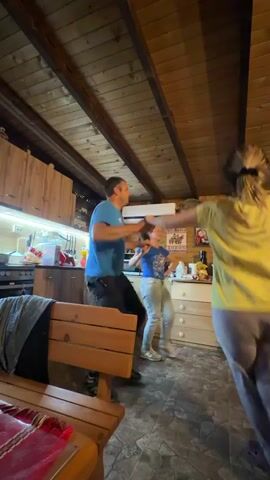 Inside a wood-paneled room that appears to be a combination living room and kitchen, a man, a woman, and a young girl are dancing energetically. The man wears a blue t-shirt and dark pants, while the woman is dressed in a yellow t-shirt and gray sweatpants. The girl wears a blue t-shirt and light gray jeans. All three face forward, moving their arms and legs in a lively, somewhat chaotic rhythm.

To the left, there is a wooden bench, table, and a glimpse of the kitchen area, which includes wooden cabinets and a countertop with various items. On the wall behind them is a flatscreen television displaying a fireplace. The wooden ceiling is supported by exposed beams. The floor is tiled with a busy pattern. The room is illuminated by overhead lighting, creating a warm and cozy atmosphere.